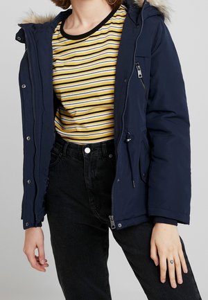 Osoba ubrana w granatową parkę na żółtą, białą i czarną pasiastą koszulę, czarne, wysokie spodnie jeansowe oraz z małym tatuażem serca na palcu.