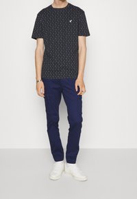 Man som bär en svart t-shirt med prickar, marinblå cargobyxor med fickor, vita sneakers och ett silverfärgat armbandsur, stojande mot en enfärgad vit bakgrund.