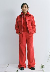 Giacca di velluto a coste rossa con due tasche sul petto, abbinata a pantaloni a coste a vita alta dello stesso materiale. Scarpe nere rotonde completano il look.