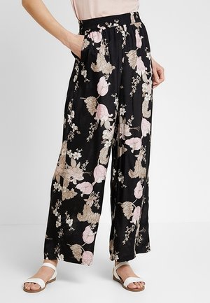 Mujer con pantalones negros de pierna ancha con estampado floral en rosa y beige, cintura elástica y sandalias blancas, de pie con una mano en el bolsillo.