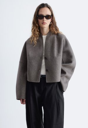 Femme aux cheveux ondulés portant une veste en laine marron oversize, des lunettes de soleil noires, une chemise blanche et un pantalon large noir sur un fond uni.