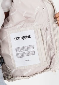 Beżowa kurtka z ociepleniem, wnętrze z powtarzającym się białym logo "SIXTH JUNE", detal tekstury oraz metka z instrukcją pielęgnacji wszyta wewnątrz.