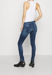 Jeans ajustados de denim azul oscuro con una textura suave, que presentan una cintura alta, bolsillos traseros y cremalleras en los tobillos, combinados con una camiseta sin mangas blanca.