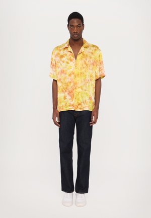 Short-sleeve, button-up shirt in yellow hues with orange patterns, paired with dark blue jeans and white sneakers. Smooth fabric texture.