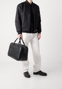 Sac de voyage en cuir noir avec double poignée, fermeture éclair et détails métalliques discrets. Associé à une veste bomber noire et un pantalon clair.
