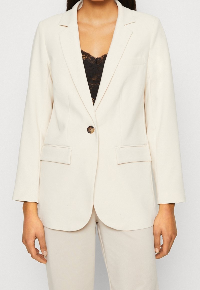 Femme portant un blazer ajusté écru à bouton unique, avec des poches à rabat, et un haut en dentelle noire en dessous, debout devant un fond uni.