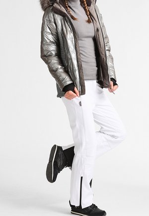 Personne portant une veste matelassée argentée avec capuche en fourrure, col roulé gris, pantalon blanc et baskets noires, debout avec une jambe pliée.