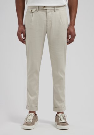 Man draagt beige cropped broek, wit overhemd en grijze suède sneakers met witte veters, staand tegen een effen achtergrond.
