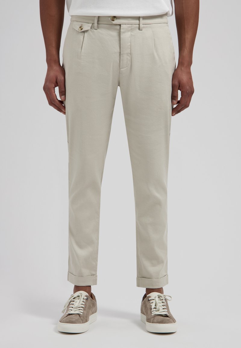 Homme portant un pantalon beige raccourci, une chemise blanche et des baskets en daim gris avec des lacets blancs, debout devant un fond uni.