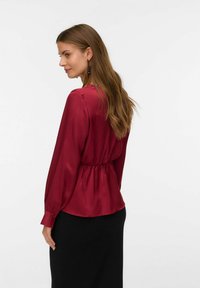 Rubinrød satinbluse med lange ærmer, snøret talje og knapdetaljer på manchetterne, båret med en sort nederdel. Glat tekstur.