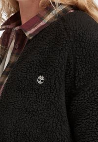 Czarna kurtka z polaru o teksturowanej powierzchni z haftowanym logo. Pod nią widoczna jest koszula w kratę w kolorach bordowym i brązowym.