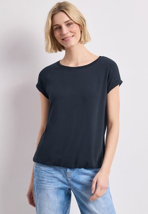 Uśmiechnięta kobieta o krótkich blond włosach, ubrana w granatowy top z krótkim rękawem i jasnoniebieskie dżinsy na jednolitym tle.