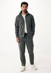 Grijze ritsjack met hoge kraag, gecombineerd met bijpassende grijze broek. Witte t-shirt eronder en witte sneakers. Zacht, textuurstof.