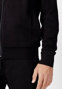Sweatshirt zippé noir avec une texture douce, des poignets et un ourlet côtelés, et des poches latérales, présentant un design ajusté et un style minimaliste.