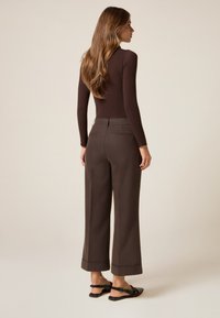 Top de manga larga marrón combinado con pantalones marrones de pierna ancha con dobladillos. Modelo también lleva sandalias planas negras, visto desde la parte de atrás.