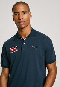 Navy poloshirt met een zachte textuur, voorzien van een geborduurde Britse vlag op de borst en een wit logo. Korte mouwen en een klassieke kraag.