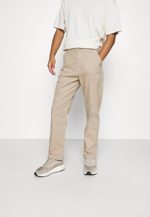 Man wearing beige trousers, white t-shirt, and grey sneakers standing with one hand in pocket against a plain background.