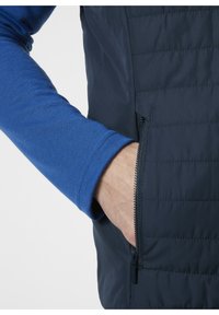 Granatowa pikowana kamizelka z zamkiem błyskawicznym, połączona z niebieską koszulą z długim rękawem. Posiada fakturowany materiał i kontrastowe detale szwów.