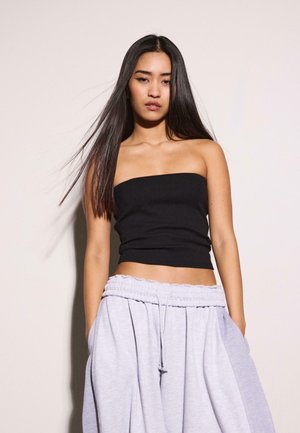 Mlada ženska z dolgimi ravnimi temnimi lasmi, oblečena v črn zgornji del brez naramnic in široke svetlo sive trenirke, stoji ob navadni ozadju.