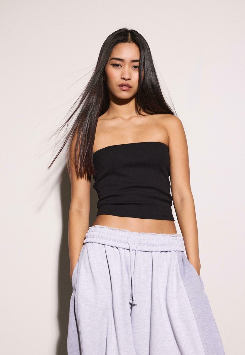 Ung kvinde med langt, glat mørkt hår iført en sort stropfri top og løse, lysegrå joggingbukser, stående mod en ensfarvet baggrund.