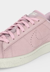 Ανοιχτόροζ αθλητικό παπούτσι Nike με υφή σουέντ, ροζ κορδόνια που ταιριάζουν, διάτρητο σχέδιο swoosh και λευκή υφή σόλας.