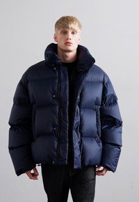 Junger Mann in einer übergroßen dunkelblauen Daunenjacke mit hohem Kragen, der vor einem schlichten hellen Hintergrund steht.