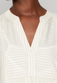 Blouse beige clair avec des rayures verticales subtiles, décolleté en V, texture douce et coupe décontractée. Présente un design de empiècement à l'avant.