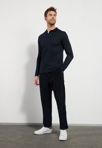 Marineblå langærmet polo-shirt med vertikale hvide striber, der har krave og en to-knaps placket, parret med mørke kordfløjlsbukser og hvide sneakers.