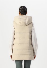 Personne avec des cheveux tressés portant un gilet matelassé beige à capuche sur une chemise claire et un pantalon sombre, de dos contre un fond uni.