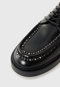 Black leather shoe features a rounded toe, studded accents along the edge, and a textured sole with a smooth finish. Laced design.