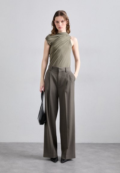 Haut drapé vert olive avec un design d'épaule asymétrique, associé à un pantalon marron à jambes larges. Modèle portant un sac à main noir et des chaussures pointues.