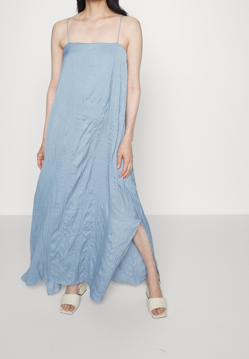 Femme portant une robe maxi sans manches bleu clair avec de fines bretelles et une fente latérale, associée à des sandales blanches à talons blocs et bouts ouverts.