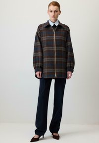 Karirana jakna z zadrgo spredaj, v odtenkih modre in rjave, iz mehke tkanine, z obesnimi rameni in prevelikim krojem, kombinirana s temno modrimi hlačami.