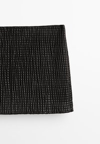 Mini-jupe noire texturée avec un motif matelassé et un léger éclat, présentée sur un fond blanc.