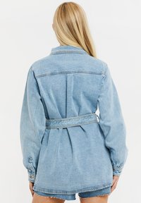 Chaqueta de mezclilla azul claro con cintura ajustada por cinturón, mangas largas y puños abotonados. Presenta un cuello clásico y un pliegue en la parte trasera.