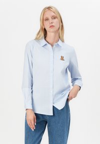 Ljusblå och vitrandig skjorta med knappar, prydd med en broderad teddybjörn på vänster sida, i kombination med blå denimjeans.