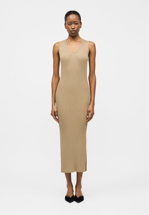 Žena stojaca čelom vpred, oblečená v béžových midi šatách bez rukávov z rebrového pleteného materiálu s výstrihom do V a čiernymi topánkami so špicatou špičkou.