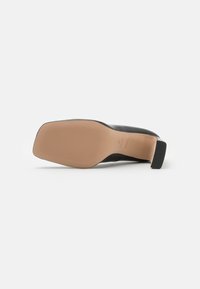 Semelle d'une chaussure à talon en cuir noir avec un bout carré et une semelle antidérapante texturée beige, vue par le dessous sur un fond blanc.