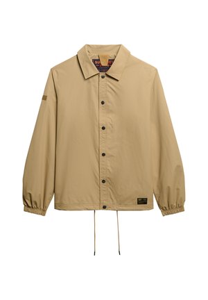 Veste beige à boutons avec un col, des poignets élastiques, un ourlet à cordon et un patch logo sur le bas à gauche. Texture de tissu lisse.