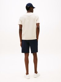 Beige cotton t-shirt, navy shorts, white sneakers, and navy cap with adjustable strap; view from behind against a light background.