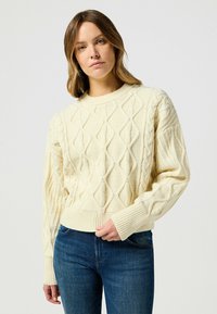 Maglione crema a maglia cable con scollatura rotonda, modelli a diamante testurizzati e polsini a costine, abbinato a jeans blu.