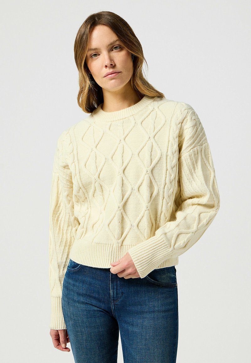 Maglione crema a maglia cable con scollatura rotonda, modelli a diamante testurizzati e polsini a costine, abbinato a jeans blu.