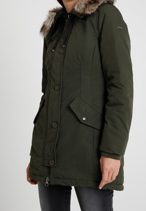 Femme portant un manteau d'hiver vert foncé avec capuche bordée de fourrure, fermeture à bouton et à glissière, poches à rabat, et jean noir.