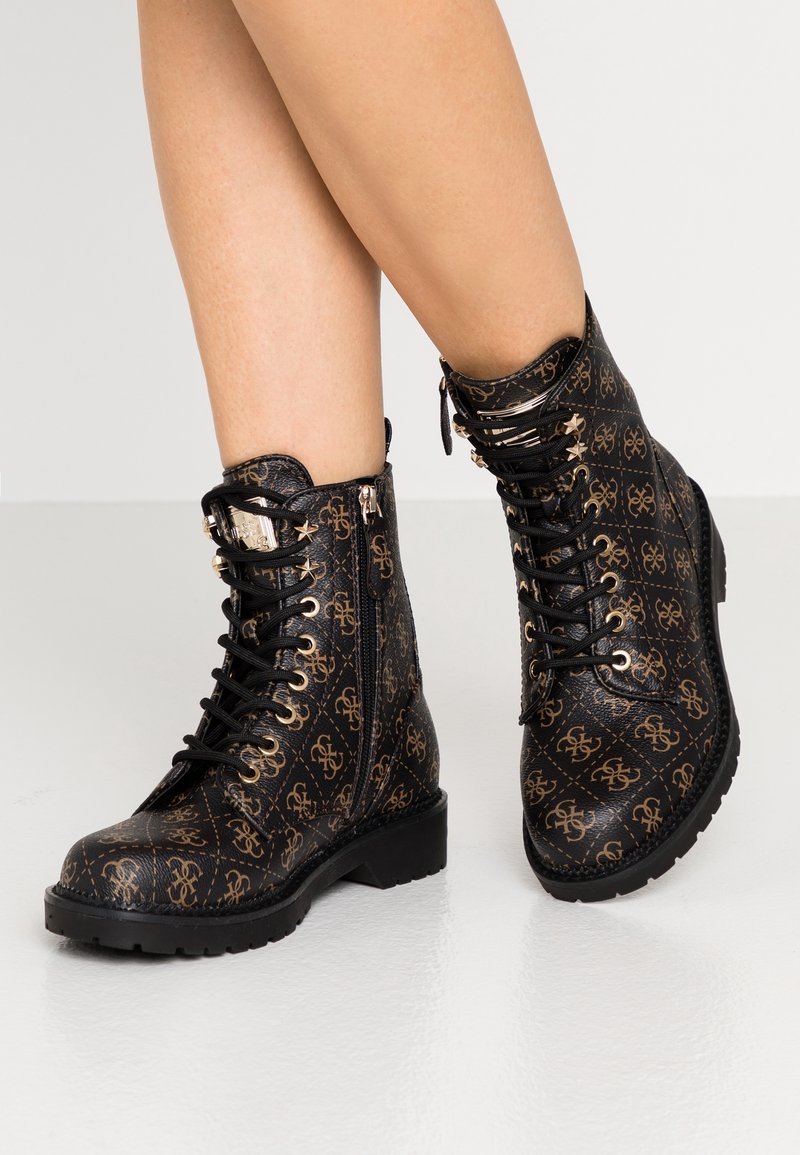 Jambes portant des bottines noires avec motif de logo doré, œillets en forme d’étoile, fermetures éclair latérales et semelles épaisses sur un sol clair.