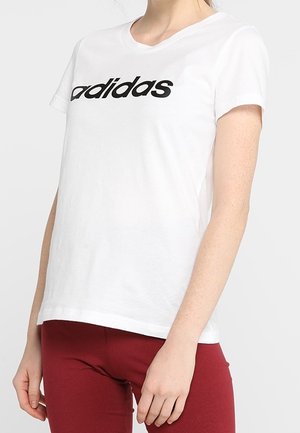 Persona che indossa una t-shirt bianca Adidas con testo del logo nero e pantaloni rossi, in piedi su uno sfondo chiaro uniforme.