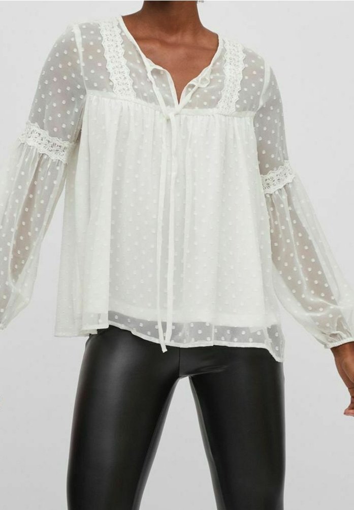 Personne portant un chemisier blanc transparent à pois avec bordure en dentelle et col à nouer, associé à un pantalon en cuir noir, sur fond uni.