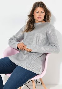 Suéter plateado de manga larga acanalado con un diseño texturizado, combinado con jeans ajustados de color azul oscuro, usado mientras está sentado en una silla rosa.