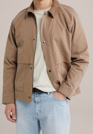 Mann trägt eine hellbraune Jacke mit Druckknöpfen über einem weißen Hemd und hellblauen Jeans, eine Hand in der Tasche.