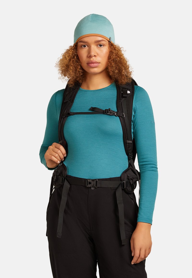 Young person with curly hair wearing a teal long-sleeve shirt and cap, adjusting black hiking backpack straps, dressed for outdoor activity.