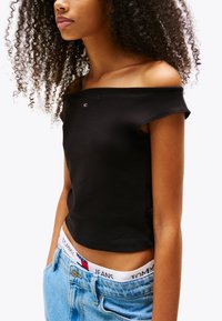 Fekete, váll nélküli crop top sima textúrával, kis logó részlettel. Magas derekú világoskék farmernadrággal párosítva.